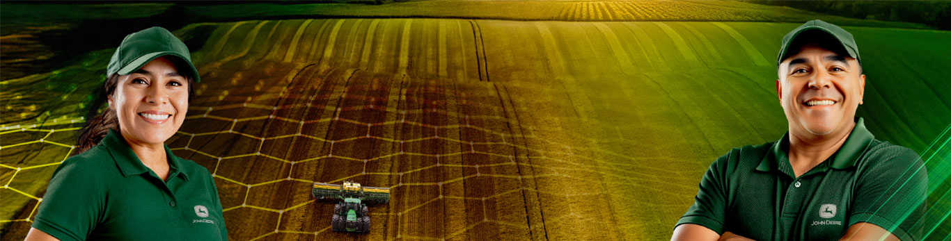 Uma mulher e um homem, ambos vestindo boné e camisa verdes, um campo com uma máquina John Deere ao fundo.