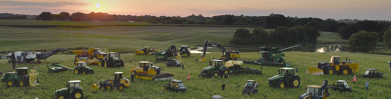 Pessoas caminhando entre vários tipos de equipamentos John Deere em um grande campo.