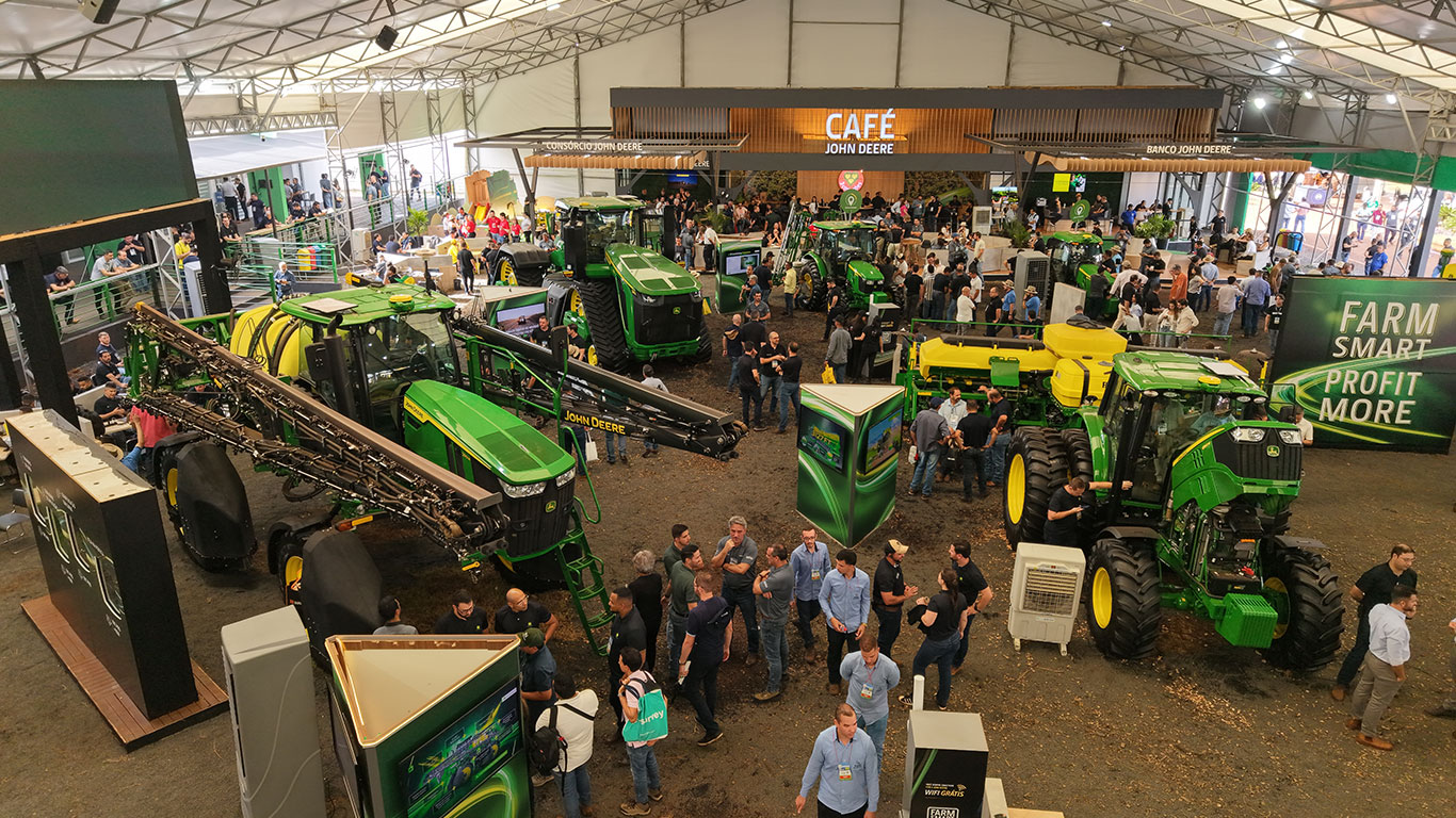 Exposição de máquinas agrícolas com pessoas, tratores e um café sob uma grande tenda.