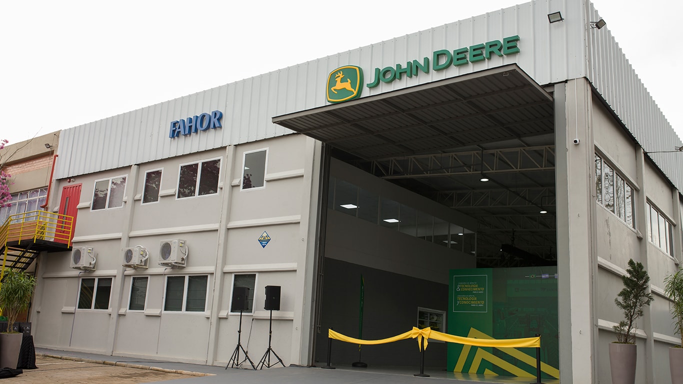 Fachada de um prédio com os logotipos da FAHOR e da John Deere. A entrada principal está aberta e decorada com uma faixa amarela, por conta da cerimônia de inauguração. Acima da entrada, o logotipo da John Deere está em destaque. 