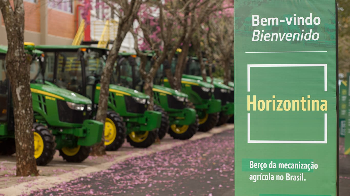 Uma fileira de tratores verdes está estacionada ao longo de uma rua arborizada com flores cor-de-rosa. Em primeiro plano, há uma placa verde com os dizeres "Bem-vindo / Bienvenido — Horizontina — Berço da mecanização agrícola no Brasil / Cuna de la mecanización agrícola en Brasil".