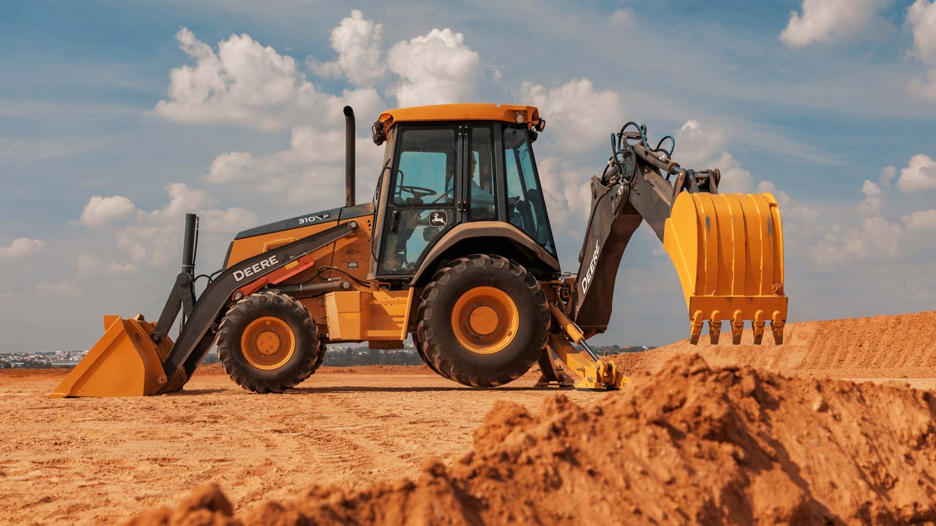 Uma retroescavadeira, modelo 310P, amarela e preta da marca John Deere está em um canteiro de obras, com montes de terra em primeiro plano. A máquina possui uma pá carregadeira na frente e um braço escavador com balde de escavação na parte traseira.
