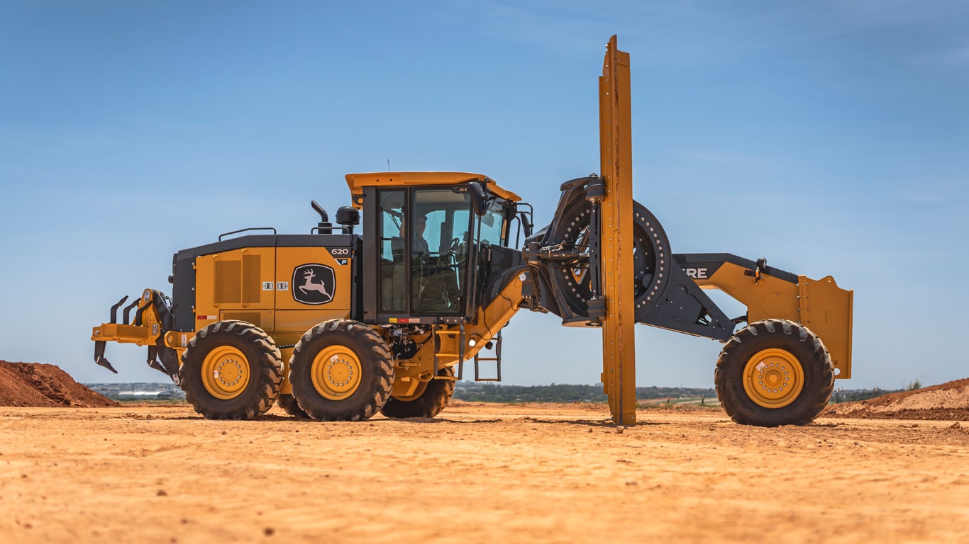 Motoniveladora John Deere 620P parada em solo arenoso, com céu claro ao fundo