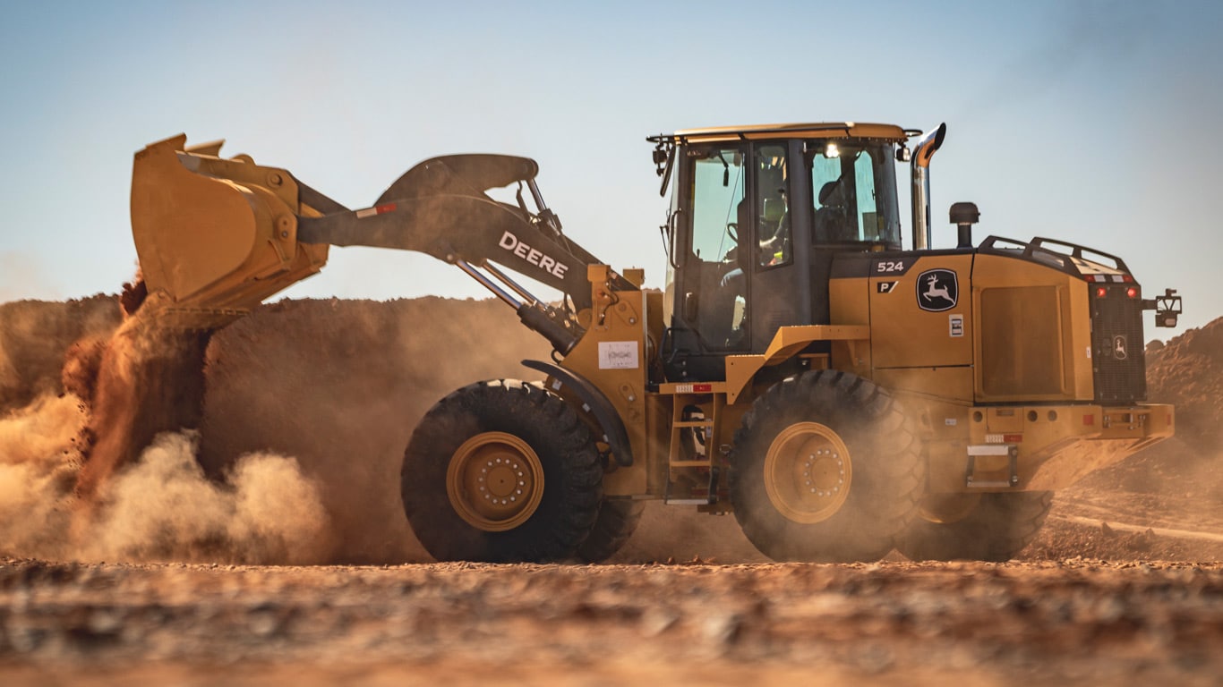 Escavadeira John Deere 524 levanta terra em solo arenoso, sob céu azul e claro