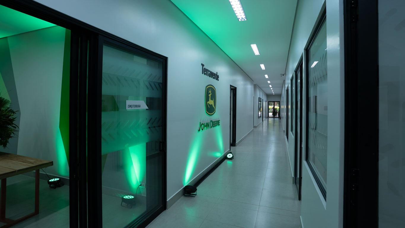 A hallway with glass-walled rooms on the left, illuminated by green lights, and a John Deere logo on the wall.