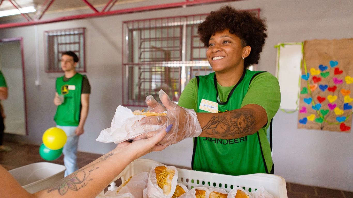 Voluntária entrega sanduíche com luvas, enquanto outras pessoas participam de atividade comunitária