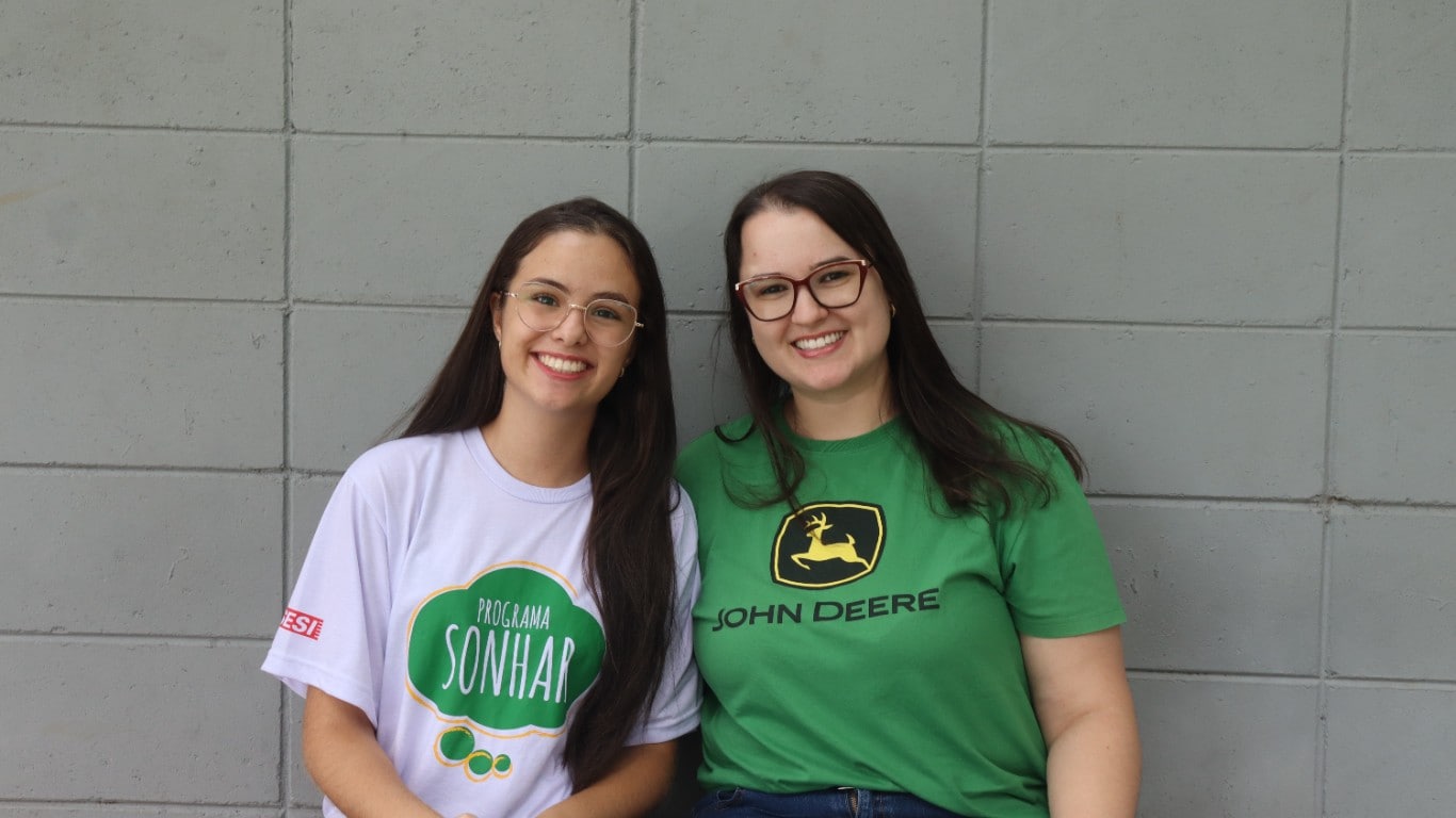 Duas pessoas sorrindo, usando camisetas de programa social, sentadas diante de parede cinza.