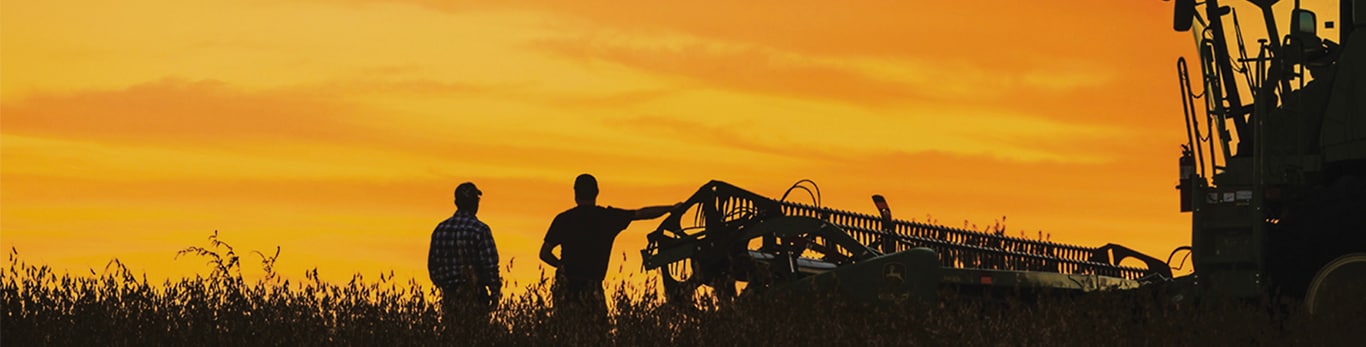 Dois agricultores em pé em um campo com uma colheitadeira John Deere ao lado deles.