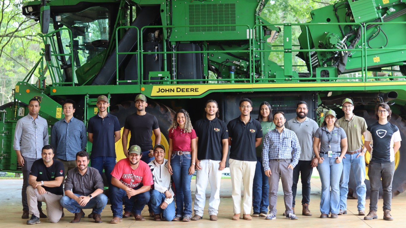 Grupo de pessoas posando em frente a máquina agrícola John Deere em ambiente industrial.