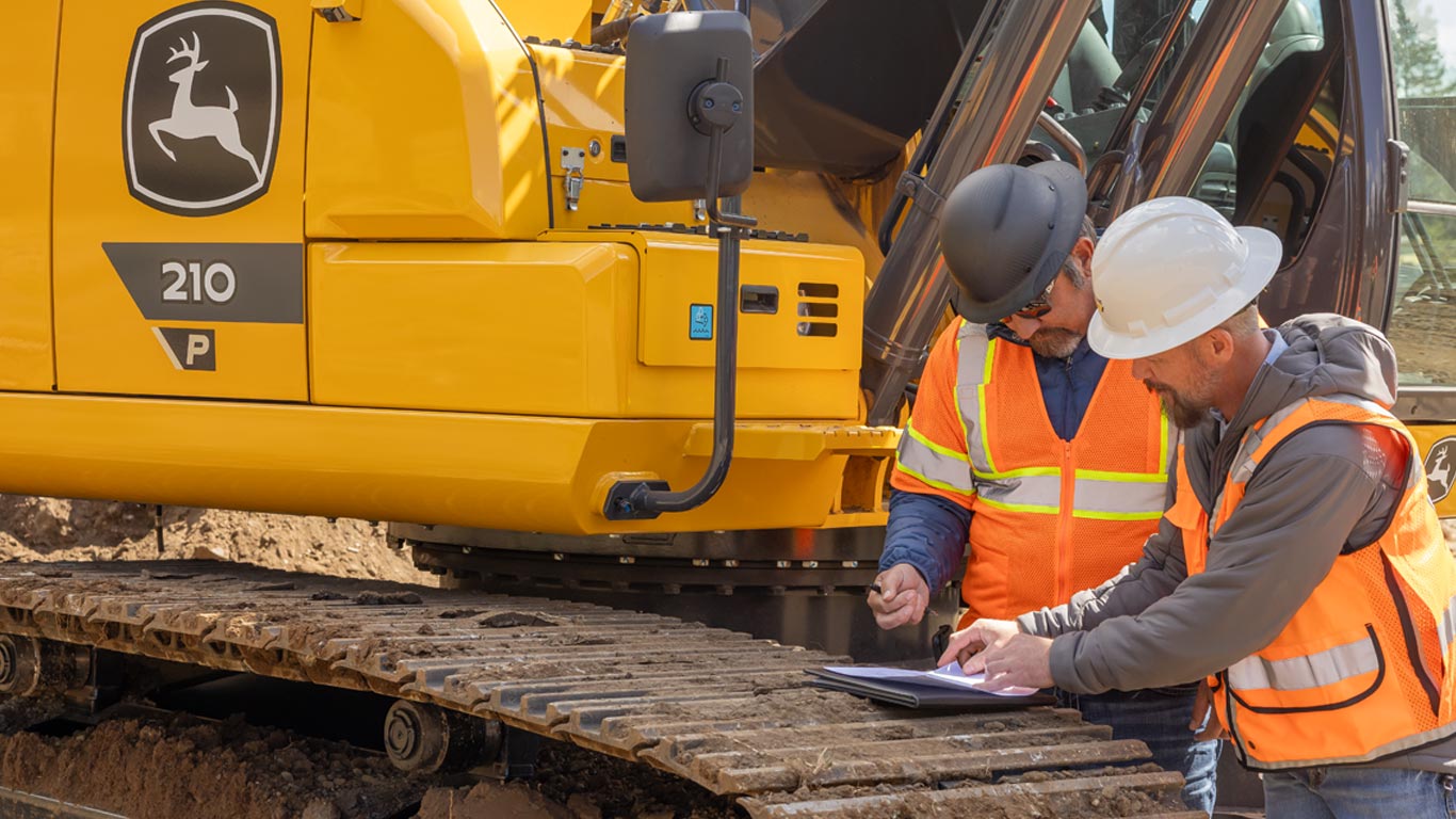 Dois trabalhadores da construção civil com equipamentos de segurança analisam documentos em uma prancheta ao lado de uma escavadeira amarela.