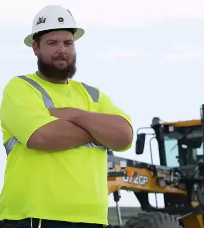 Ryan em pé na frente do equipamento para construção Deere com braços cruzados