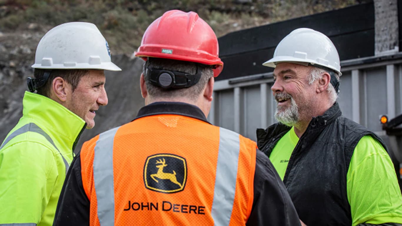 Dois homens de colete de segurança com logotipo da John Deere e capacete conversando