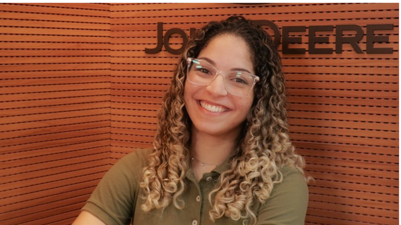 Mulher branca de cabelos enrolados e castanhos, que utiliza óculos de grau, sorri olhando para a frente enquanto está sentada, vestindo camiseta verde com o logo da John Deere