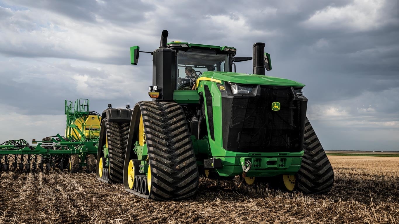 Um trator agrícola grande e moderno da marca John Deere, modelo verde com detalhes amarelos, está estacionado em um campo de solo seco e palha. O trator está acoplado a um implemento agrícola para plantio ou preparo do solo.