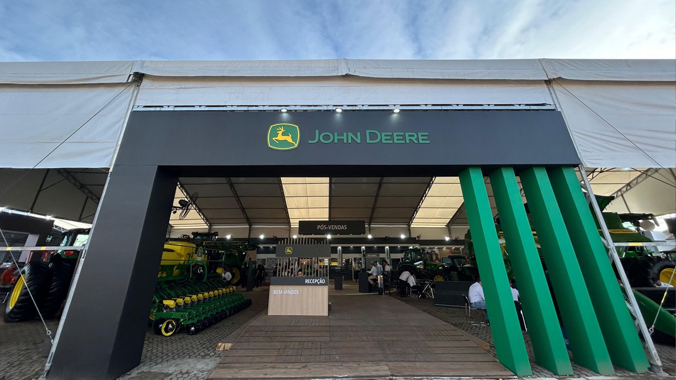 Imagem de um grande estande de exposição da John Deere em um evento ao ar livre. O nome e o logotipo da marca estão destacados no topo central da estrutura. O estande tem design moderno, com elementos verticais verdes à direita e uma entrada aberta.