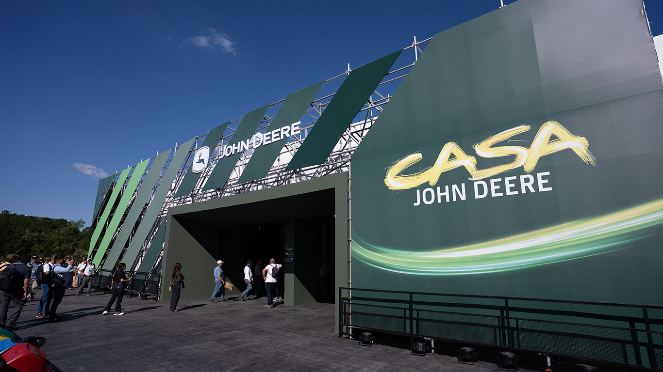 Estrutura ao ar livre da Casa John Deere, em verde, com logotipo e pessoas circulando sob céu azul.