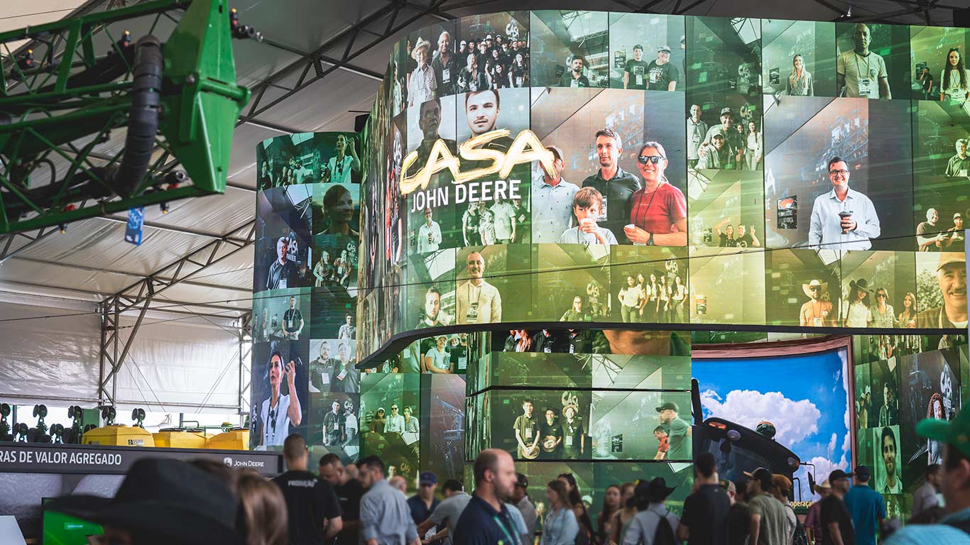 Estande da John&nbsp;Deere em feira agro, com visitantes, pain&eacute;is de LED curvos e bra&ccedil;o mec&acirc;nico verde