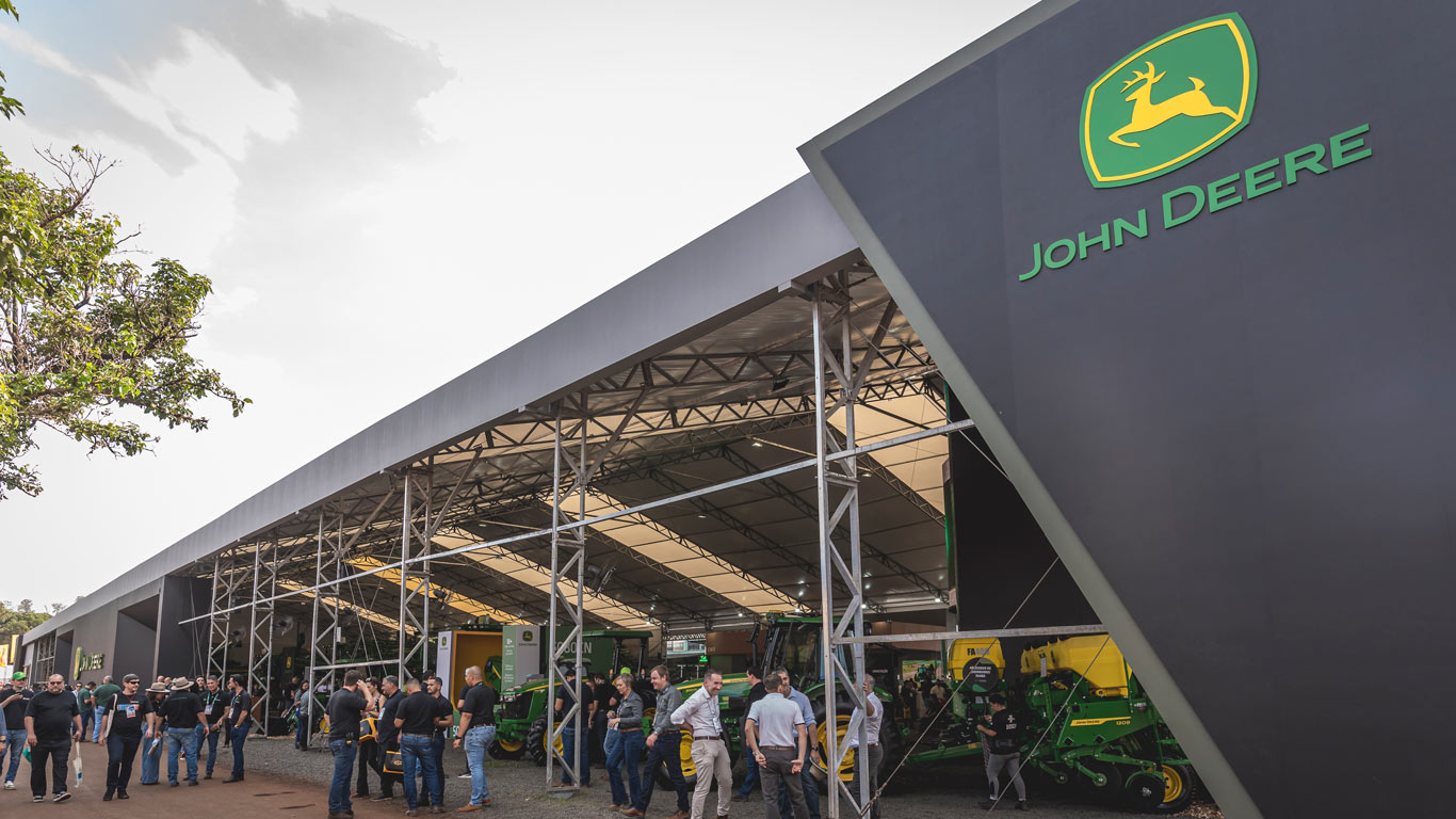 &Aacute;rea externa do estande John&nbsp;Deere em feira agro, com fachada preta, logotipo verde, m&aacute;quinas ao fundo e visitantes.
