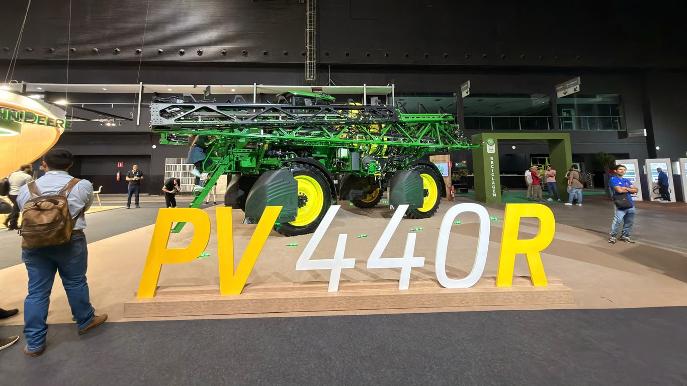 Em um estande de evento, o pulverizador agrícola John Deere PV 440R, está exposto sobre uma plataforma. À frente da máquina, há letras grandes 'PV 440R'.