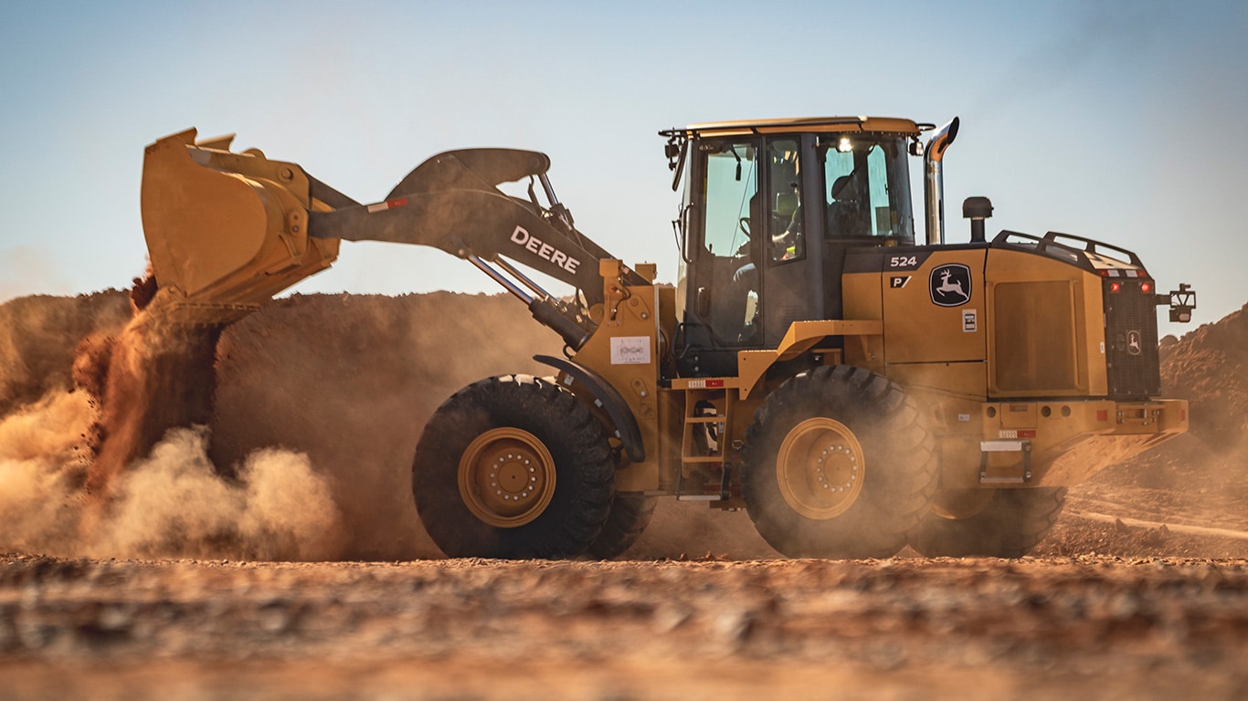 Em uma área de terraplenagem sob céu aberto, uma pá-carregadeira amarela da John Deere trabalha movimentando terra