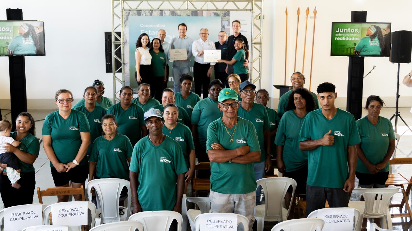 Em um sal&atilde;o iluminado, um grupo numeroso de pessoas veste camisetas verdes e est&aacute; posicionado &agrave; frente, em p&eacute;.