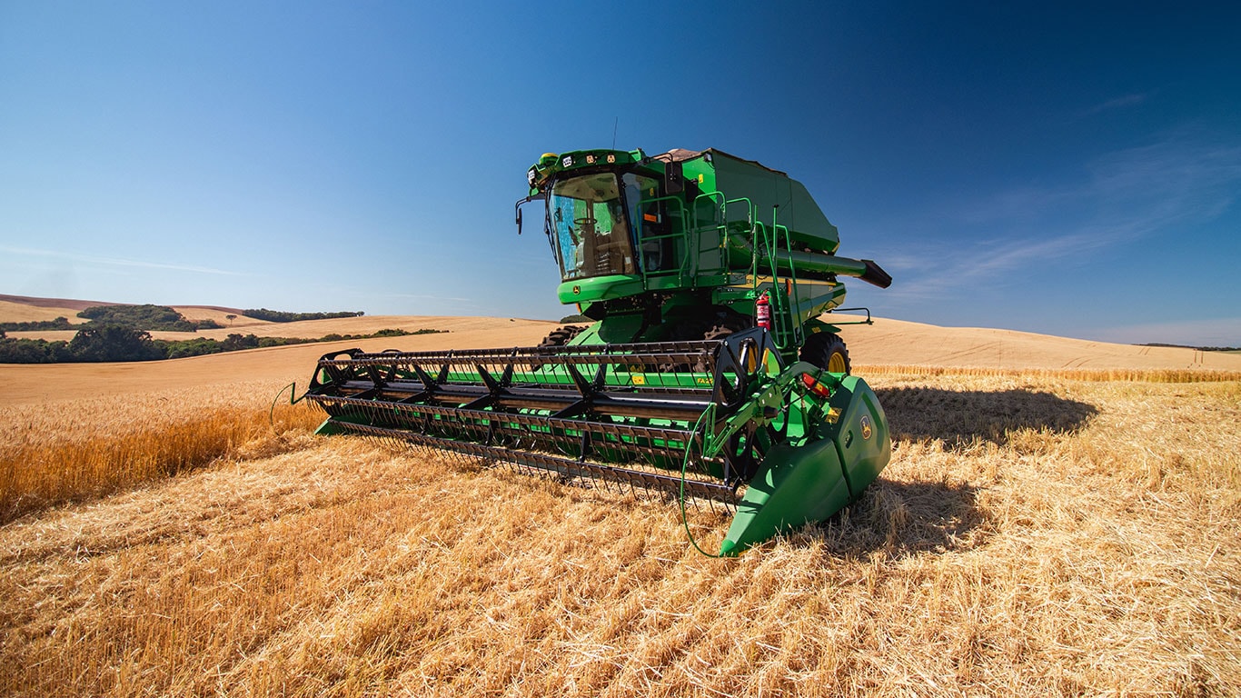 Colheitadeira John Deere S4 de frente colhendo trigo sob céu azul intenso, com paisagem rural ao fundo.