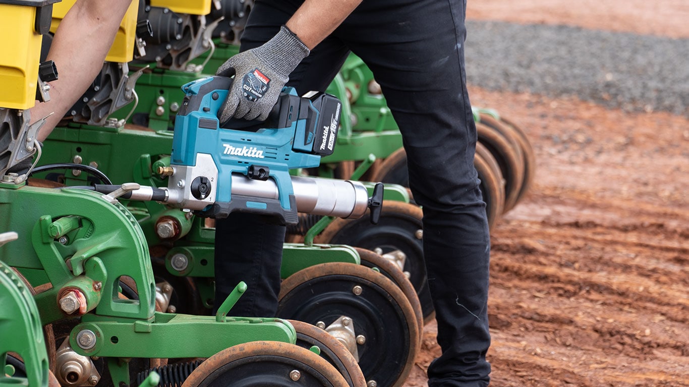Pessoa usando ferramenta el&eacute;trica Makita azul e prata para manuten&ccedil;&atilde;o em m&aacute;quina agr&iacute;cola verde, ao lado de grandes rodas met&aacute;licas sobre solo avermelhado.