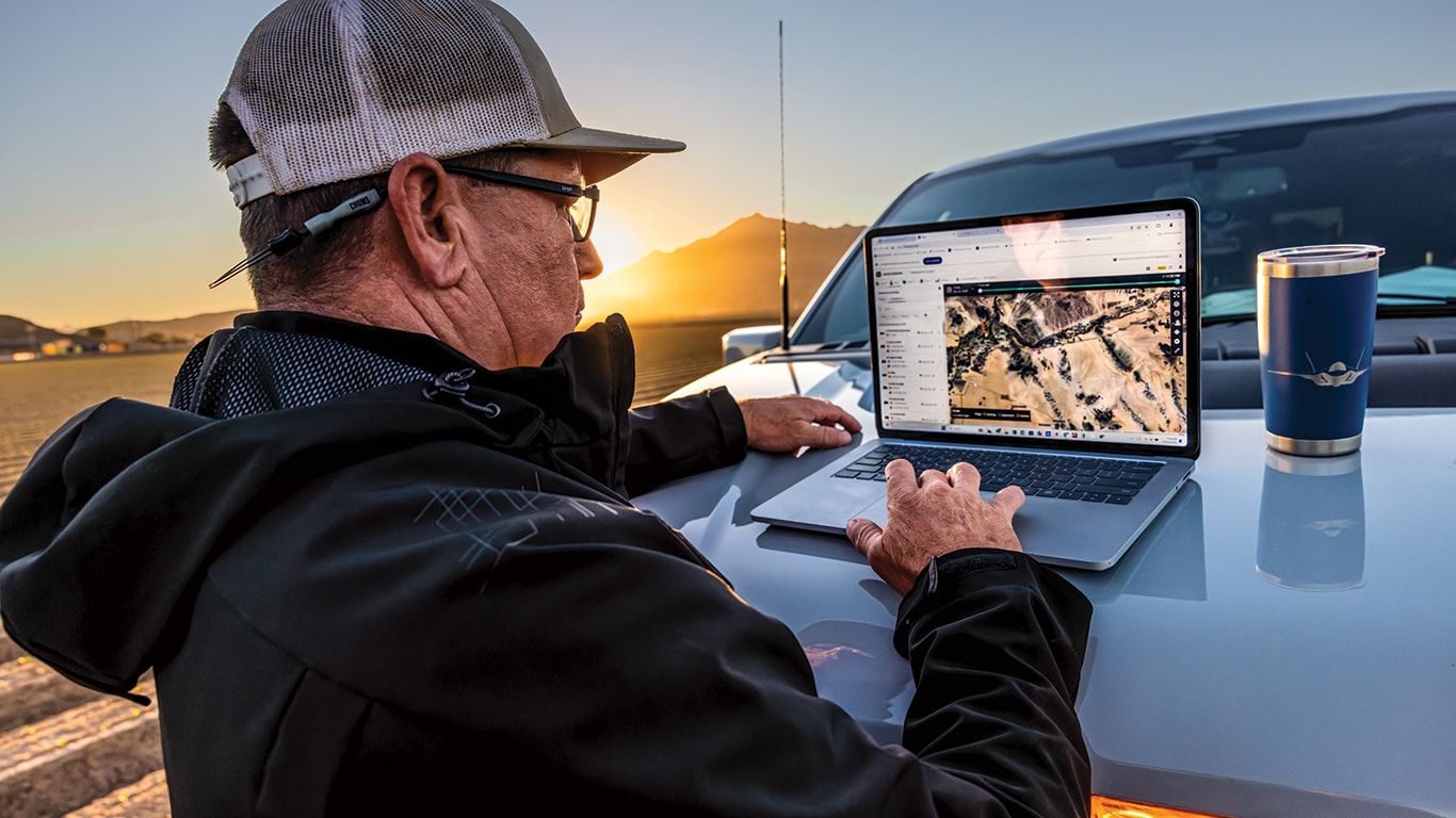 Pessoa com boné e óculos trabalha em um laptop apoiado no capô de um veículo. Na tela do computador, há uma imagem de satélite. Uma das mãos navega pelo mapa, enquanto a outra repousa sobre o carro. Ao lado do laptop, há um copo térmico com tampa.