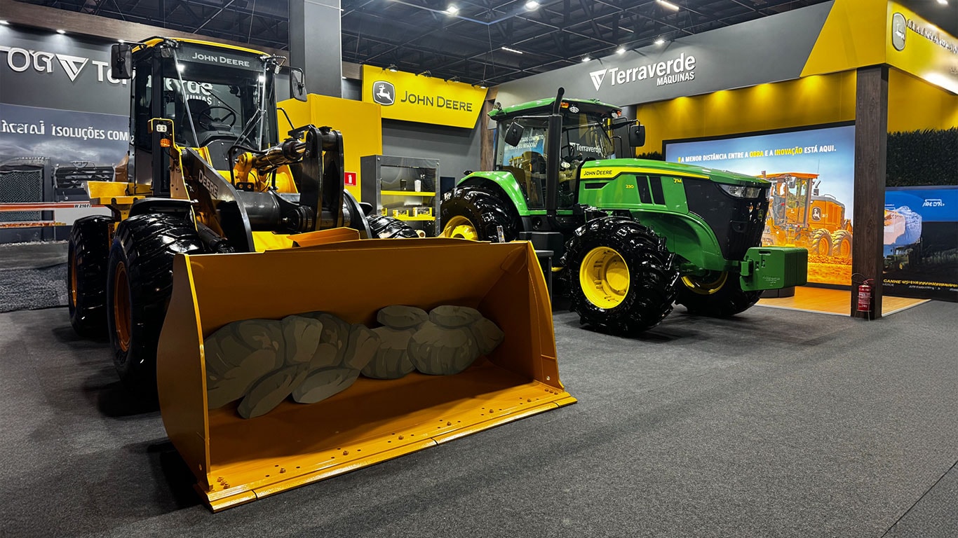 Foto de um estande da John Deere no Salão do Automóveis. À esquerda, uma pá-carregadeira amarela com caçamba grande voltada para frente. À direita, um trator verde com rodas grandes e cabine fechada. Ao fundo, painéis com logotipos John Deere e Terraverde Máquinas, iluminação intensa e piso cinza.
