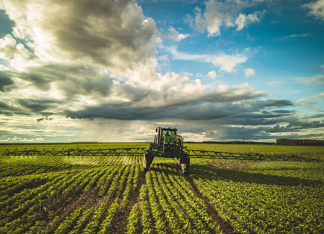 Pulverizador agrícola visto de frente com barras abertas em plantação, sob céu dramático.