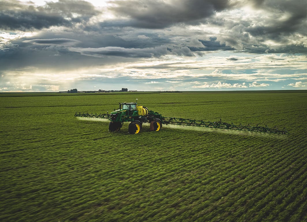 Pulverizador agrícola aplicando insumos em campo verde sob céu nublado.