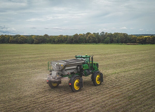 Distribuidor agrícola espalhando insumos em campo aberto sob céu nublado.