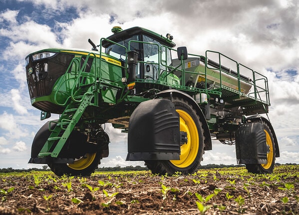 Pulverizador agrícola autopropelido verde em plantação, visto de baixo sob céu nublado.