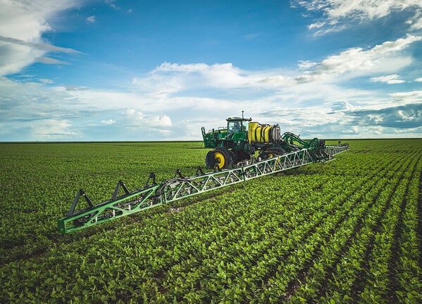 Pulverizador agrícola com barras longas aplicando insumos em lavoura verde sob céu azul.