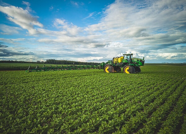 Pulverizador agrícola com barras longas operando em lavoura verde sob céu parcialmente nublado.