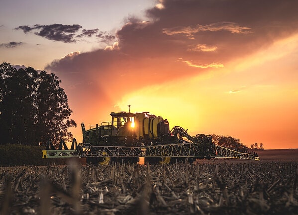 Pulverizador agrícola operando ao pôr do sol em campo colhido.