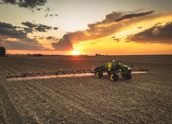 Pulverizador agrícola trabalhando ao pôr do sol em campo colhido, com barras estendidas.
