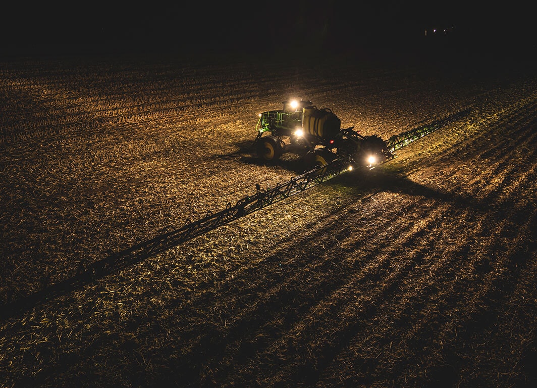 Pulverizador agrícola operando à noite com barras abertas e iluminação ativa no campo.