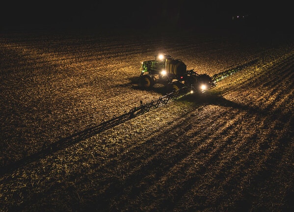 Pulverizador agrícola operando à noite com barras abertas e iluminação ativa no campo.