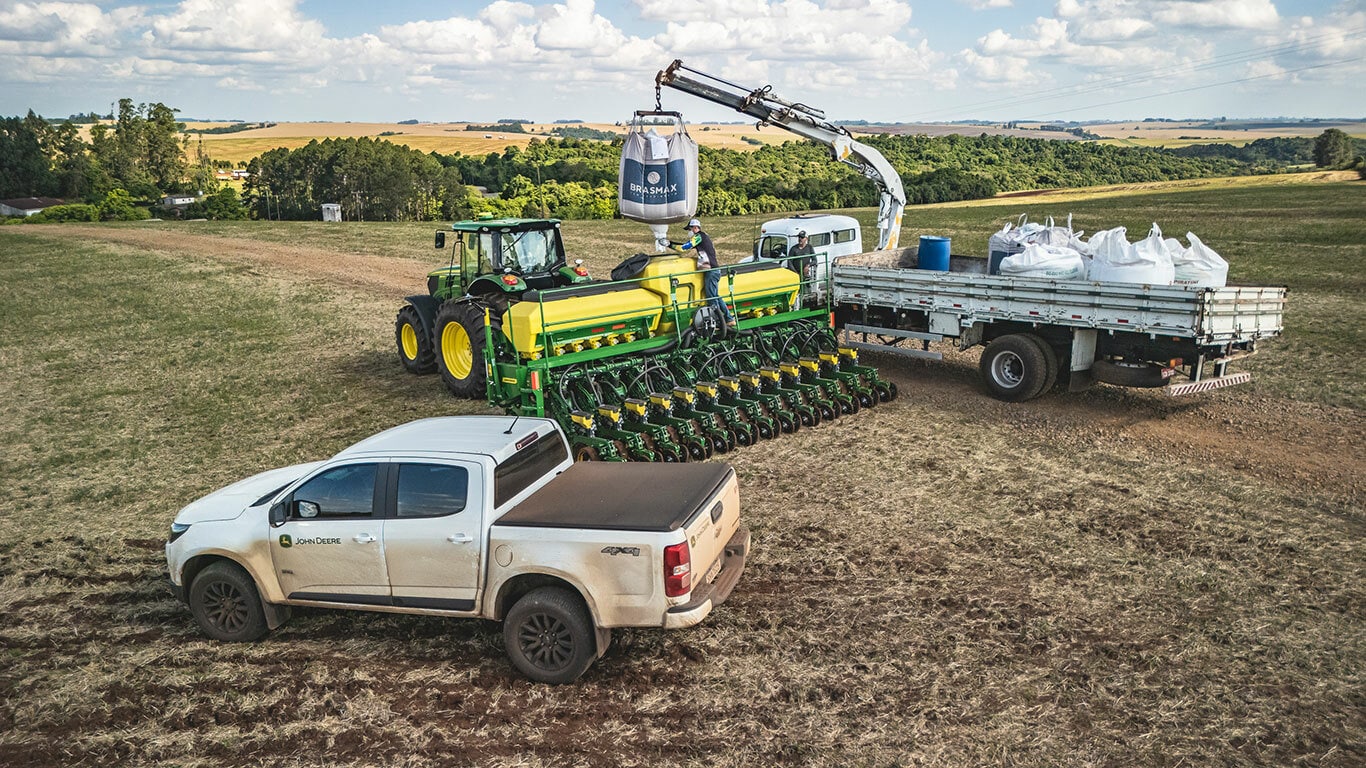 Máquina John Deere série 1200 no campo, com uma caminhonete e um caminhão.