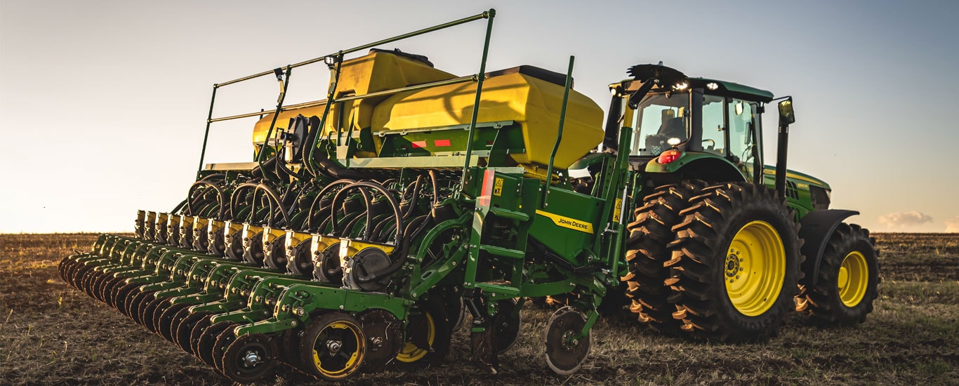 Uma plantadeira verde e amarela da John Deere presa a um trator, contra um pôr do sol sobre um campo recém-arado.
