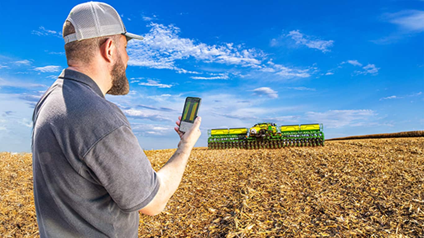 Um homem com um celular na mão, ao fundo uma plantadeira 3100 trabalhando no campo.