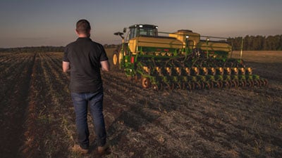 Homem a frente de uma máquina John Deere série 1200 no campo ao entardecer.