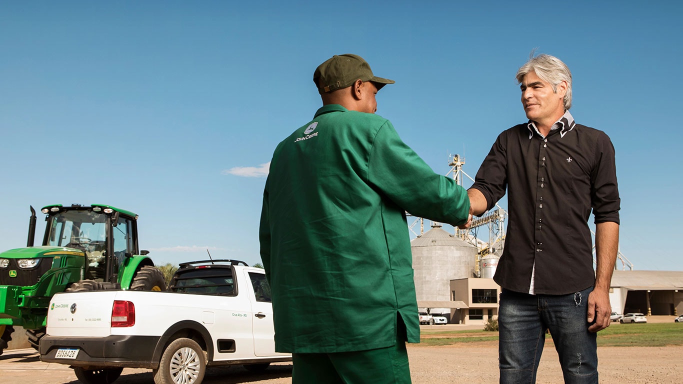 Dois homens apertando as mãos na frente de uma máquina John Deere em um campo.