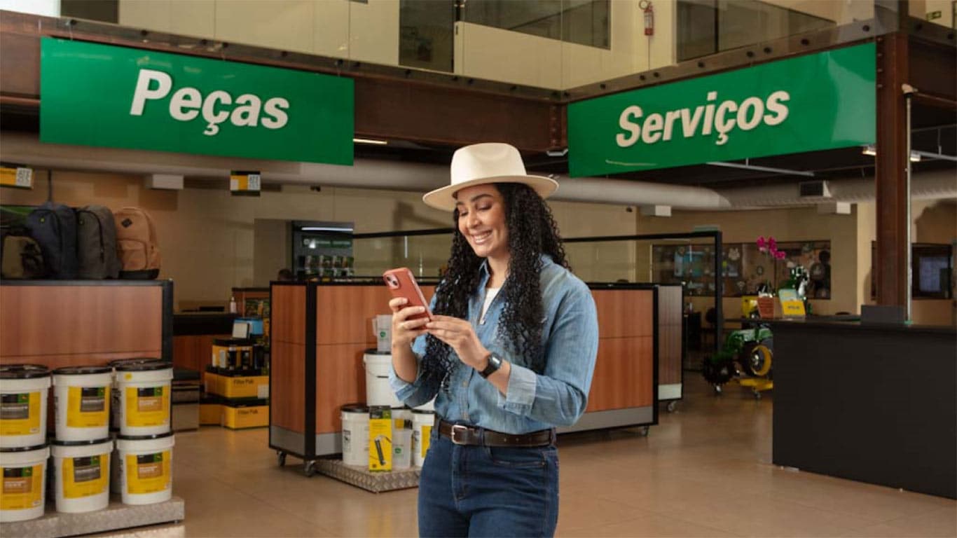 Uma mulher de cabelo longo e cacheado usando um chapéu branco, mexendo no celular na frente de um estande de Peças e Serviços.