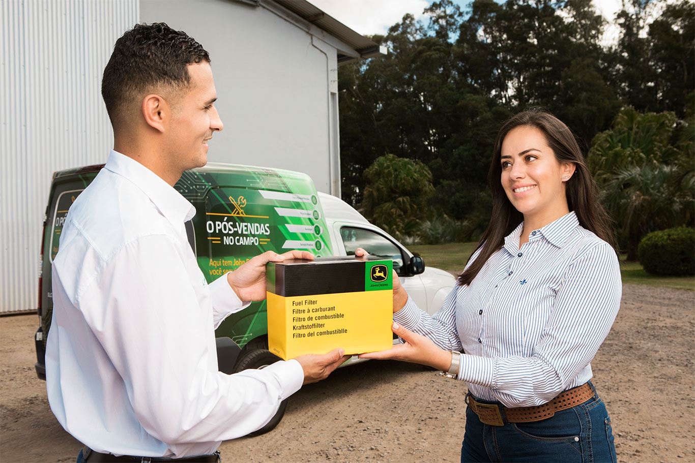 Um homem e uma mulher segurando uma caixa de filtro de combustível na frente de uma van de Pós Vendas John Deere.